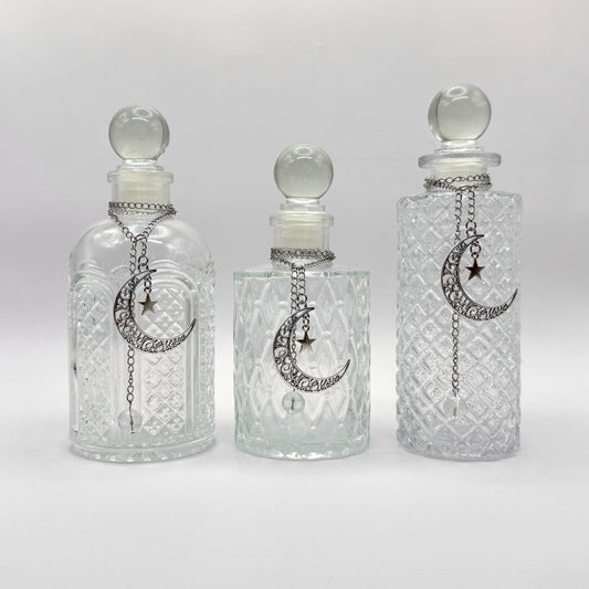 Three clear glass perfume bottles with stoppers and silver crescent moon and star chain ornaments, displayed against a light gray background.