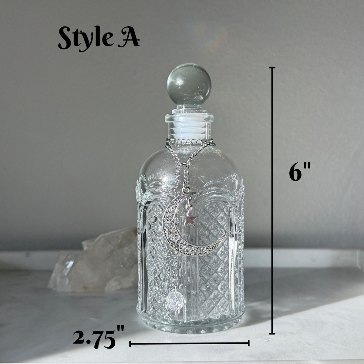 A clear glass decanter bottle with a spherical stopper and a silver-toned chain with a star pendant, shown on a light surface against a gray background. Black measurement lines indicate dimensions: 6 inches tall and 2.