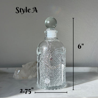 A clear glass decanter bottle with a spherical stopper and a silver-toned chain with a star pendant, shown on a light surface against a gray background. Black measurement lines indicate dimensions: 6 inches tall and 2.