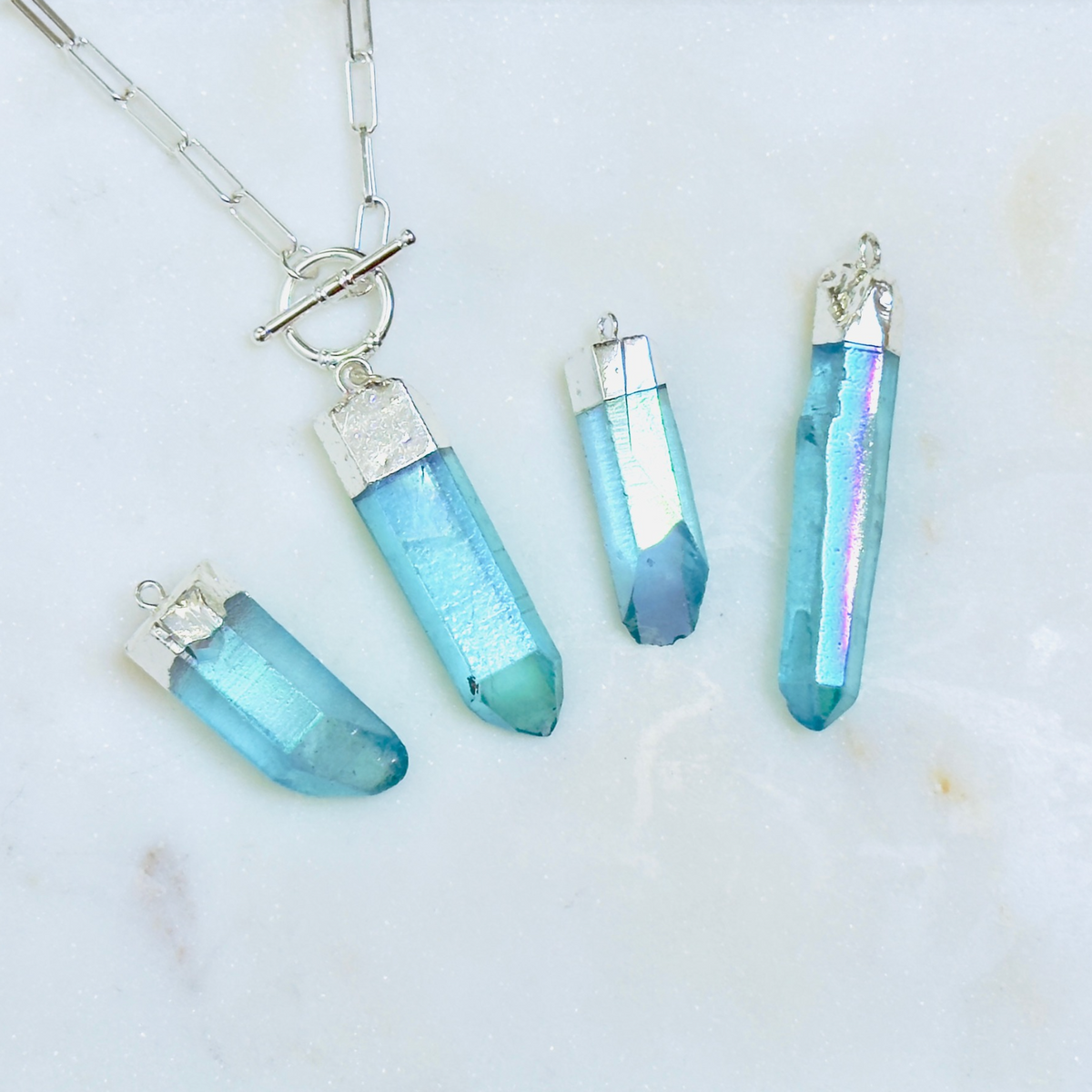Four blue iridescent crystal point pendants with silver caps and a silver chain, arranged on a white marble surface.