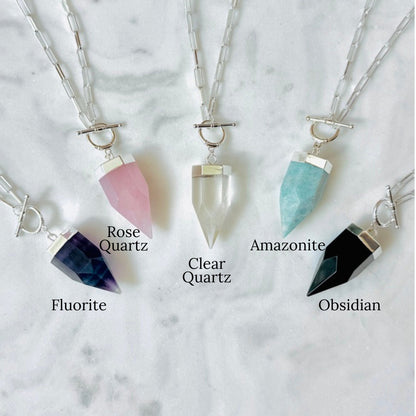 Four crystal point pendant necklaces on silver chains, labeled Fluorite, Rose Quartz, Clear Quartz, and Amazonite, arranged on a light marble background.