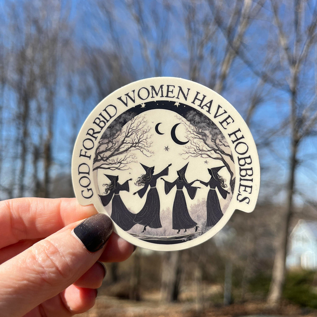 A hand with dark nail polish holds a round sticker outdoors against a blurred background of bare trees and blue sky. The sticker has a black and white illustration of three witches holding a staff under a crescent moon and stars, with the text 'GOD FORBID WOMEN HAVE HORRORS' around the edge.