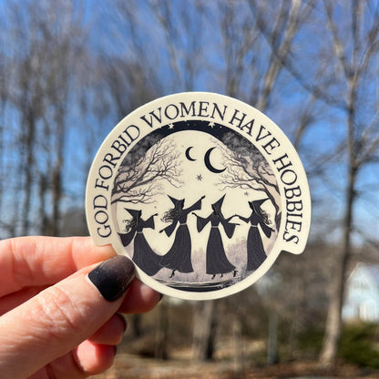 A hand with dark nail polish holds a round sticker outdoors against a blurred background of bare trees and blue sky. The sticker has a black and white illustration of three witches holding a staff under a crescent moon and stars, with the text 'GOD FORBID WOMEN HAVE HORRORS' around the edge.